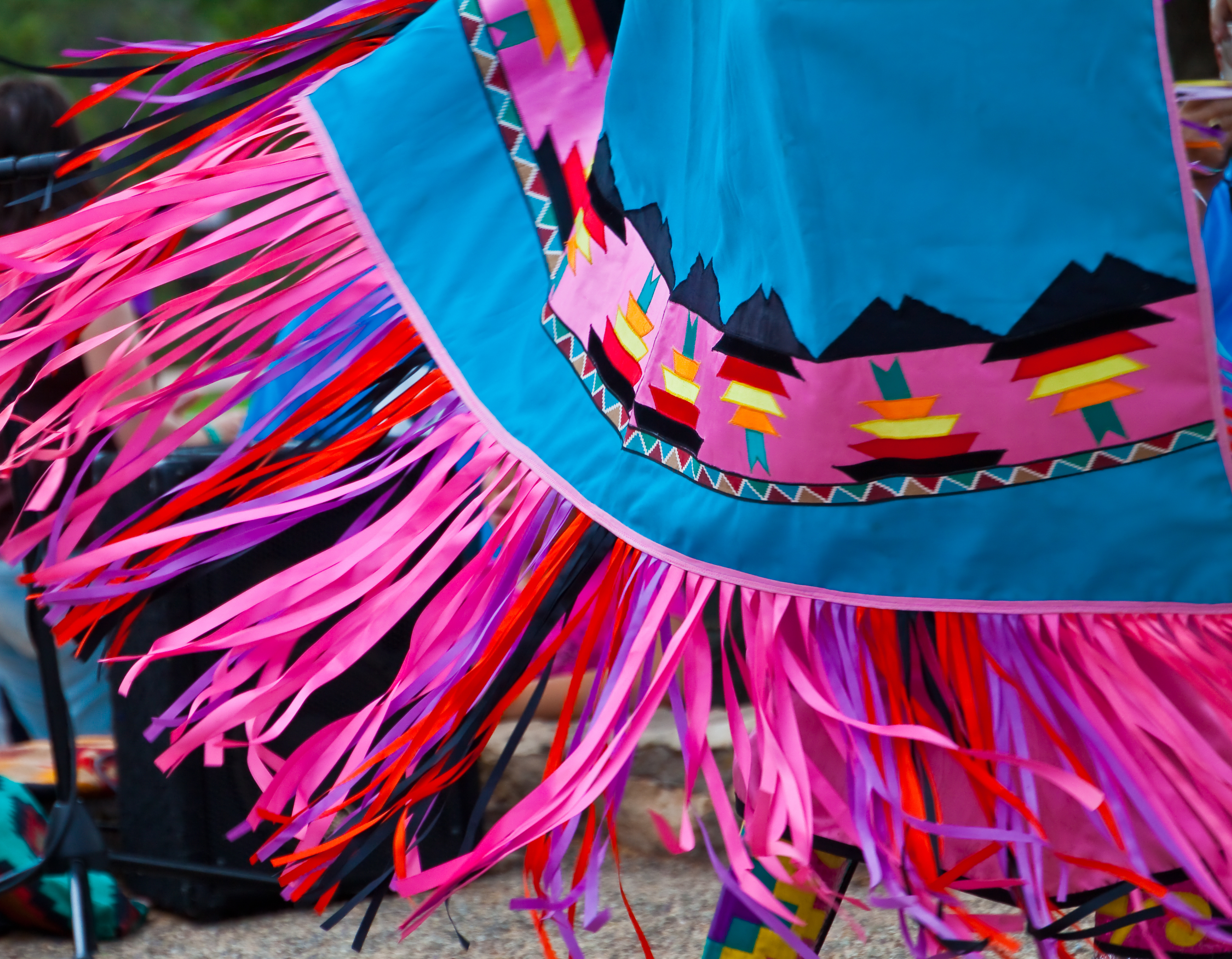 Image of portion of fancy dance shawl in movement. The shawl is bright blue with pink fringes and colorful motif appliqué
