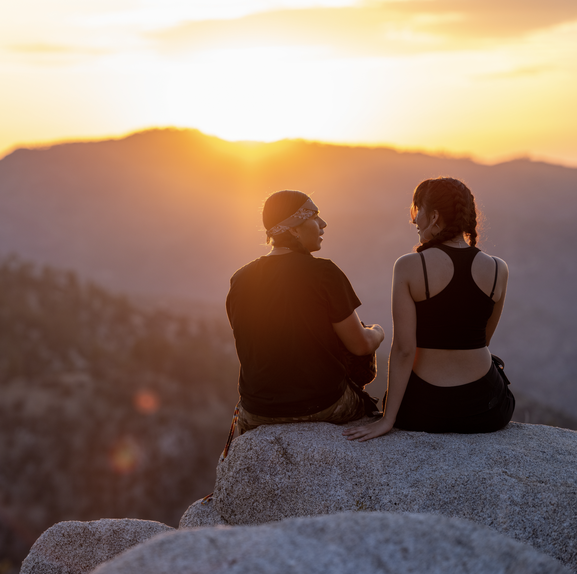 Image of two Native American people sitting on a rock ledge overlooking mountains at sunset while talking