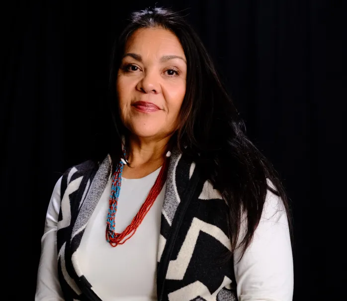 Portrait of Elaine (Juanita) Castillo wearing a black and white shirt with beaded necklace against a black background