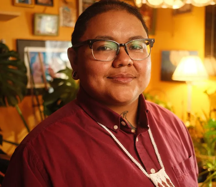 Portrait of Mimi wearing a red collared shirt and necklace in a warm background with picture frames on the wall. 